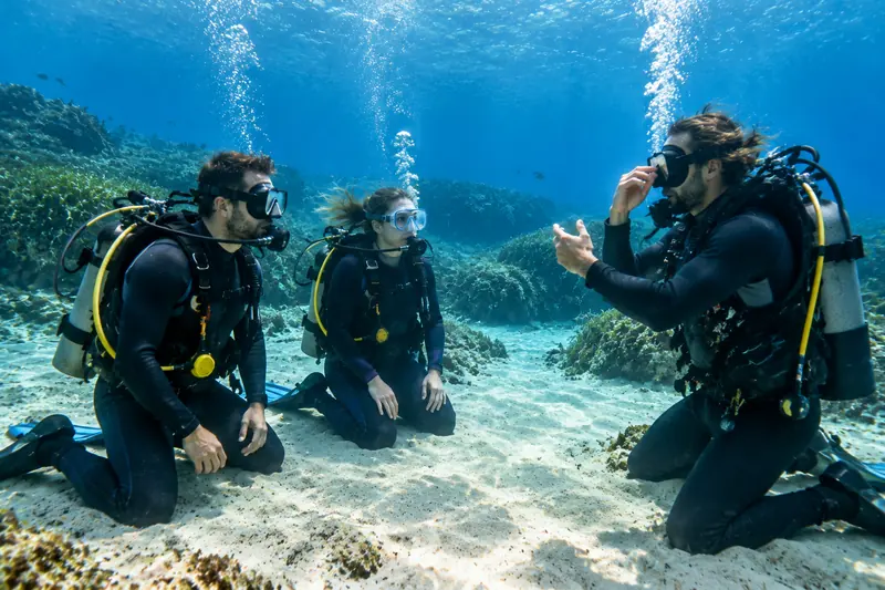 Dos alumnos de buceo practican habilidades arrodillados en la arena frente a su instructor